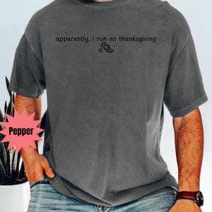 May include: Dark grey t-shirt with the text "apparently, i run on thanksgiving" and a small graphic of a running shoe. The shirt is a relaxed fit and has short sleeves. The model is wearing blue jeans and a brown watch.