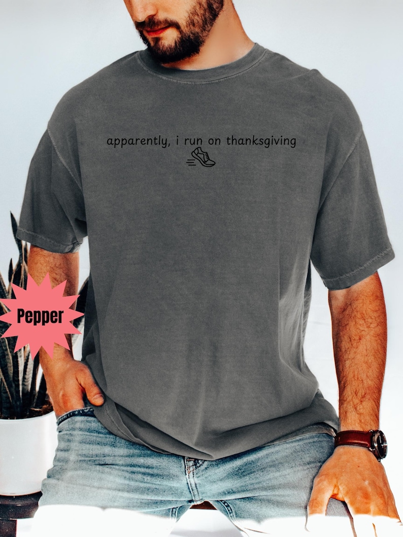 May include: Dark grey t-shirt with the text "apparently, i run on thanksgiving" and a small graphic of a running shoe. The shirt is a relaxed fit and has short sleeves. The model is wearing blue jeans and a brown watch.