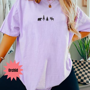 May include: Orchid-colored t-shirt with a small black graphic of a bear, two trees, and a moose. The shirt is a solid color and has short sleeves. The word "Orchid" is in a red starburst shape.