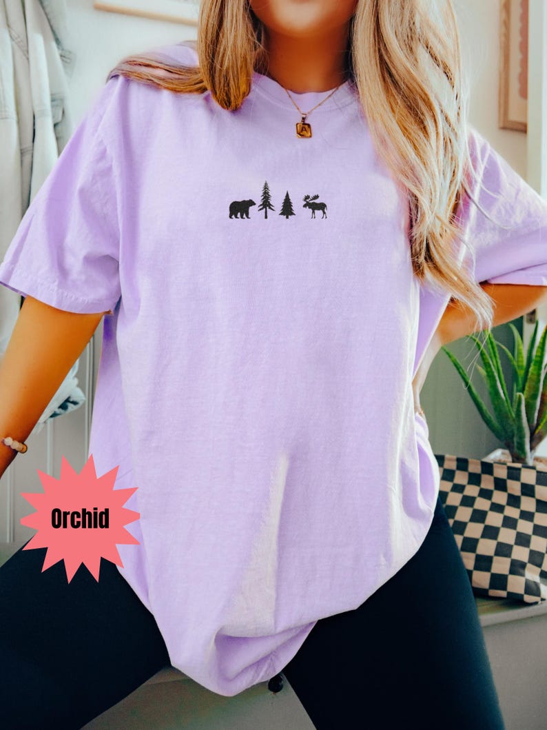 May include: Orchid-colored t-shirt with a small black graphic of a bear, two trees, and a moose. The shirt is a solid color and has short sleeves. The word "Orchid" is in a red starburst shape.
