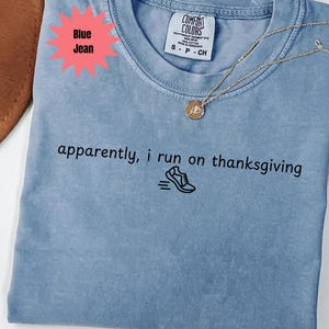 May include: A light blue t-shirt with the text "apparently, i run on thanksgiving" and a running shoe graphic. A gold necklace with a circular pendant rests on the shirt. The shirt is labeled "Blue Jean".
