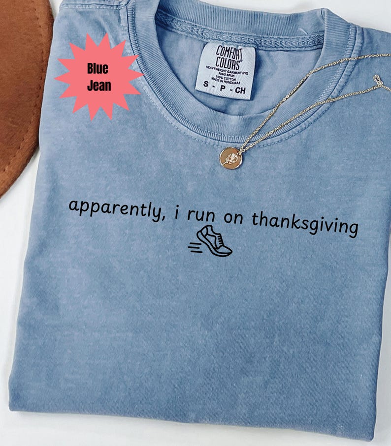 May include: A light blue t-shirt with the text "apparently, i run on thanksgiving" and a running shoe graphic. A gold necklace with a circular pendant rests on the shirt. The shirt is labeled "Blue Jean".