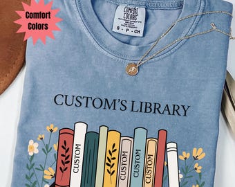 May include: A light blue Comfort Colors t-shirt with the text "CUSTOM'S LIBRARY" above a graphic of colorful books on a shelf. A gold necklace with a pendant is draped on the shirt.