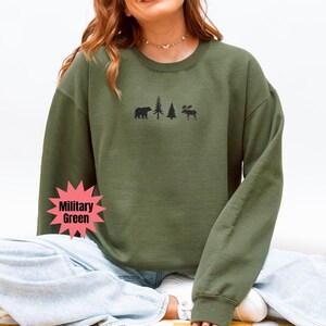 May include: Military green sweatshirt with a crew neck. The front features a black graphic of a bear, two trees, and a moose. The sweatshirt is a solid color and has long sleeves. The model is wearing blue jeans.