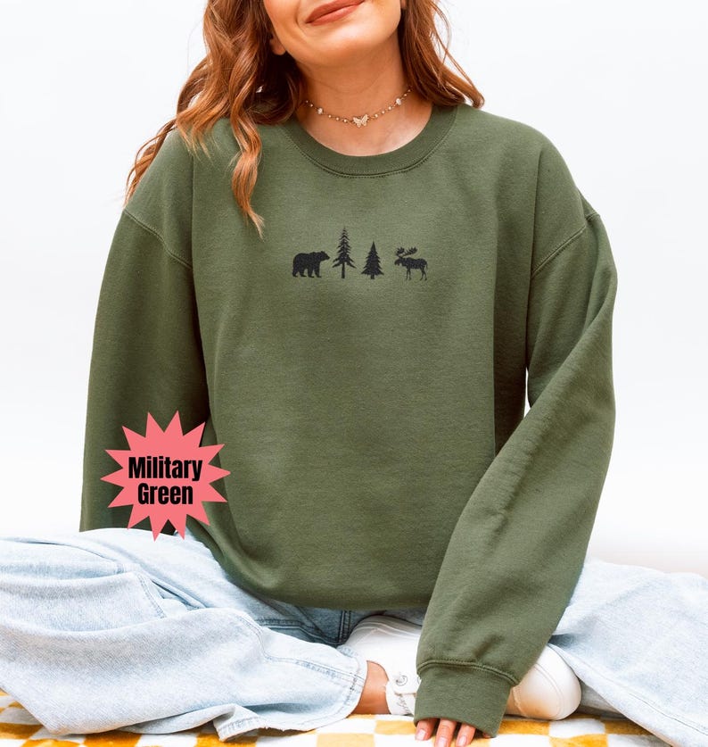 May include: Military green sweatshirt with a crew neck. The front features a black graphic of a bear, two trees, and a moose. The sweatshirt is a solid color and has long sleeves. The model is wearing blue jeans.