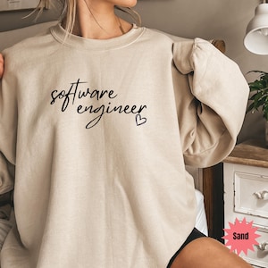 May include: A sand-coloured sweatshirt featuring the text "software engineer" in black script, with a small heart. The top has a crew neck and long sleeves.