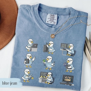 May include: Blue jean Comfort Colors t-shirt with a crew neck and short sleeves. The shirt features a graphic of a duck wearing glasses and working on computers. A gold necklace is draped across the shirt.