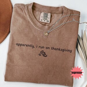 May include: Espresso-colored Comfort Colors t-shirt with the text "apparently, i run on thanksgiving" and a running shoe graphic. A silver necklace with a pendant is draped across the shirt.