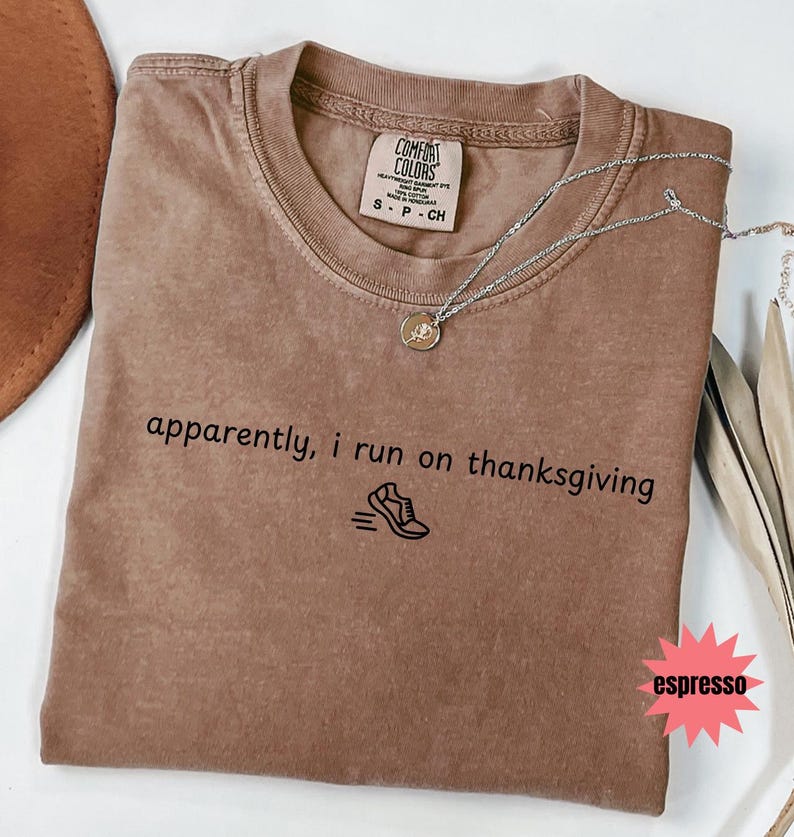 May include: Espresso-colored Comfort Colors t-shirt with the text "apparently, i run on thanksgiving" and a running shoe graphic. A silver necklace with a pendant is draped across the shirt.