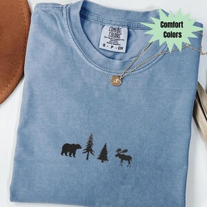 May include: A blue Comfort Colors t-shirt with a bear, trees, and moose design. The shirt has a crew neck and short sleeves. A gold necklace with a pendant is draped across the shirt.