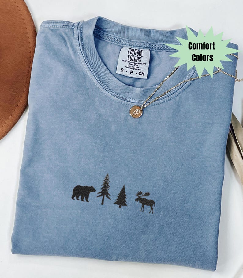 May include: A blue Comfort Colors t-shirt with a bear, trees, and moose design. The shirt has a crew neck and short sleeves. A gold necklace with a pendant is draped across the shirt.