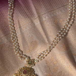 May include: A multi-strand necklace featuring small white pearls and a gold-toned pendant. The pendant is circular with a central teardrop-shaped red stone, surrounded by clear crystals and small dangling pearls at the bottom edge.