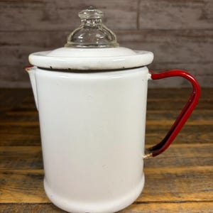 May include: White enamel coffee pot with a clear glass lid and a red handle. The vintage pot has a cylindrical body and a spout. The handle is a bright red color. The pot is sitting on a wooden surface.
