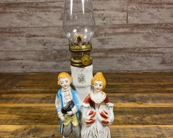 May include: A vintage oil lamp with a clear glass chimney and a decorative base featuring a couple in period clothing. The base is white with gold accents, and the figures are painted in vibrant colors.