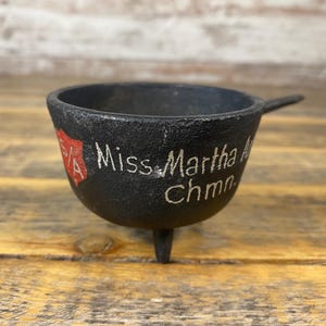 May include: A small, black cast iron bowl with three legs and a handle. The bowl has the words "Miss Martha A Chmn" in white lettering on the side, along with a red and white emblem. The bowl sits on a wooden surface.