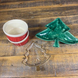 Op de afbeelding: Een verzameling kerstservies. Inclusief twee rode en witte ramekins, een groene keramische kerstboomvormige schaal en een heldere glazen boomvormige schaal met gouden rand. De schalen staan op een houten oppervlak.