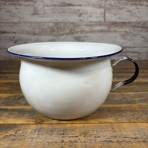 May include: A white enamel chamber pot with a smooth, rounded basin and a wide, flared rim. The rim is accented with a thin, dark blue border. A single, curved black handle is attached to the side, providing a grip for this vintage-style vessel.