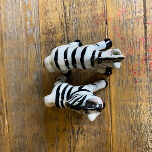 May include: Two ceramic zebra figurines, one black and white zebra is standing on its hind legs, the other is lying down. Both zebras have black and white stripes.