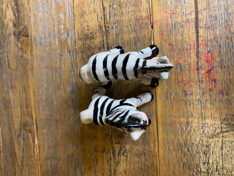 May include: Two ceramic zebra figurines, one black and white zebra is standing on its hind legs, the other is lying down. Both zebras have black and white stripes.