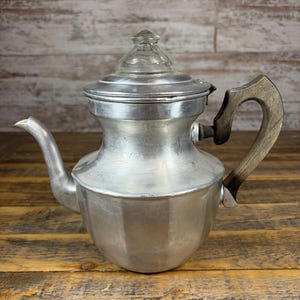 Könnte beinhalten: Eine silberfarbene Kaffeekanne aus Metall mit gebogenem Ausguss, einem dunkelbraunen Griff in Holzoptik und einem klaren Glasknauf auf dem Deckel. Der Körper der Kanne hat eine abgerundete Basis, die sich nach oben hin verjüngt.