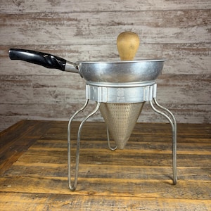 May include: A vintage aluminium food mill with a conical perforated strainer, three metal legs, a black plastic handle, and a wooden pestle. The metal band features embossed text including the words Foley and Made in USA.