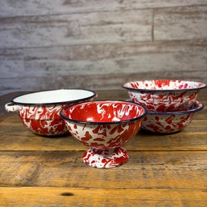 May include: A collection of enamelware bowls with a red and white speckled pattern. The bowls have a black rim and a white interior. One bowl is on a pedestal base. The bowls are on a wooden surface.