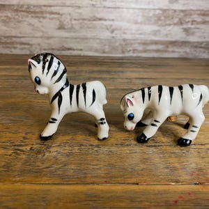May include: Two small ceramic zebra figurines. The zebras are white with black stripes and have black eyes. The zebras are standing on a wooden surface.