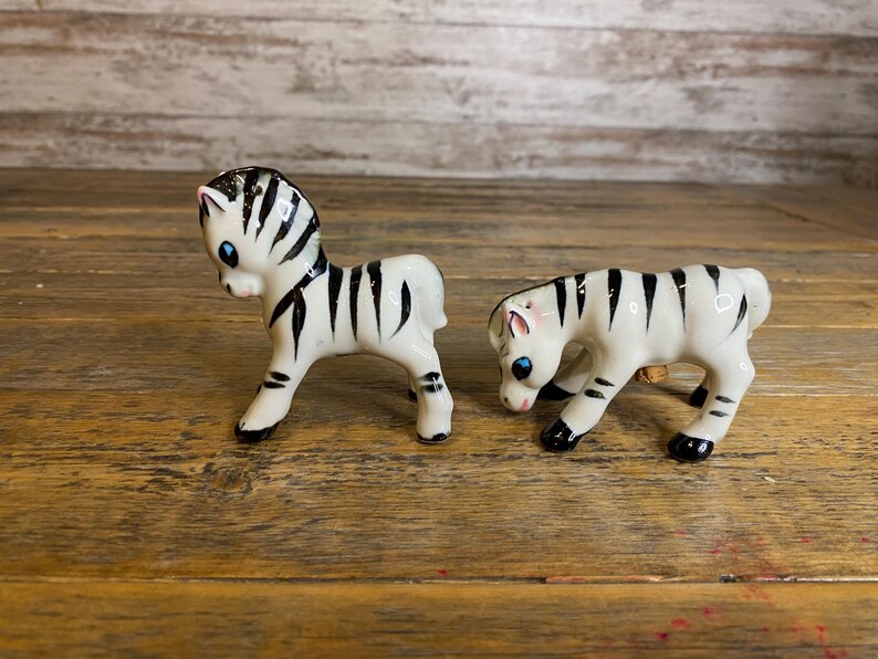 May include: Two small ceramic zebra figurines. The zebras are white with black stripes and have black eyes. The zebras are standing on a wooden surface.