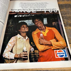 K&ouml;nnte beinhalten: Vintage Pepsi-Werbung mit einem l&auml;chelnden Paar, das Pepsi-Flaschen h&auml;lt. Der Mann tr&auml;gt ein orangefarbenes Hemd, die Frau ein Oberteil mit Blumenmuster. Das Pepsi-Logo ist zu sehen, zusammen mit dem Text "You've got a lot to live. Pepsi's got a lot to give."