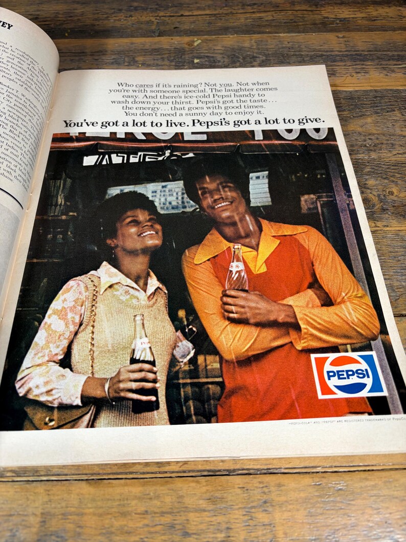 K&ouml;nnte beinhalten: Vintage Pepsi-Werbung mit einem l&auml;chelnden Paar, das Pepsi-Flaschen h&auml;lt. Der Mann tr&auml;gt ein orangefarbenes Hemd, die Frau ein Oberteil mit Blumenmuster. Das Pepsi-Logo ist zu sehen, zusammen mit dem Text "You've got a lot to live. Pepsi's got a lot to give."