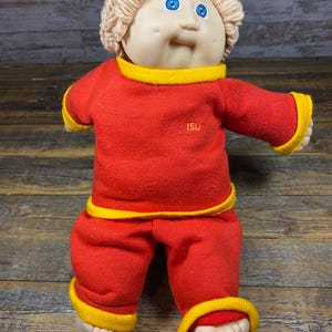 May include: A Cabbage Patch Kid doll wearing a red and yellow outfit with the letters "ISU" embroidered on the front. The doll has blonde yarn hair and blue eyes.