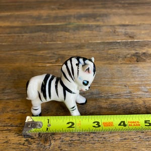 May include: A small ceramic zebra figurine with black and white stripes and blue eyes. The figurine is about 2 inches tall and is standing on a wooden surface.