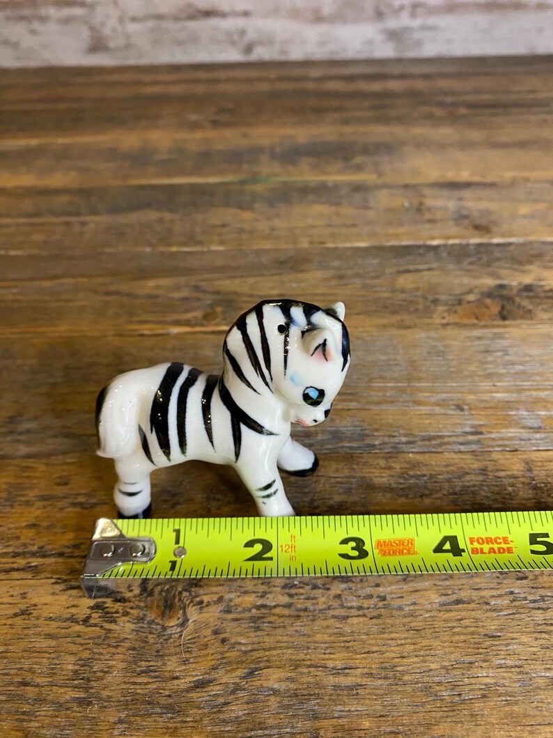 May include: A small ceramic zebra figurine with black and white stripes and blue eyes. The figurine is about 2 inches tall and is standing on a wooden surface.