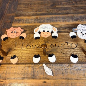 May include: A rectangular wooden wall hook rack featuring three painted animal heads: a pig, a cow, and a sheep. The board is inscribed with the text "I Love Country" and includes three dark brown wooden pegs for hanging items, along with painted hooves.