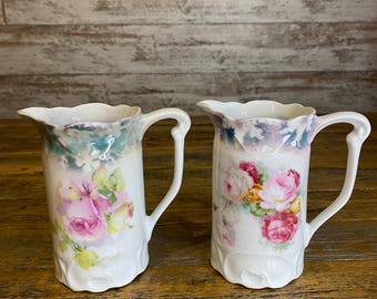 May include: Two white ceramic pitchers with floral designs. The pitchers have a scalloped rim and a handle. The floral designs are in shades of pink, yellow, and green.