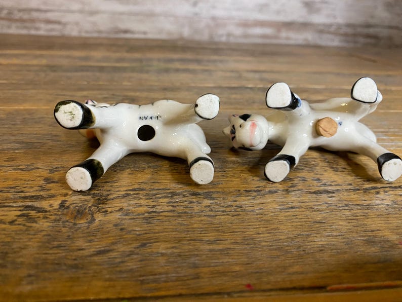 May include: A pair of black and white ceramic salt and pepper shakers shaped like cows. The shakers are upside down, revealing the openings for salt and pepper. The shakers are marked "Japan" on the bottom.