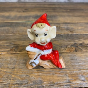 May include: A ceramic figurine of a red-hatted elf sitting on a log. The elf has a white face with painted features and is wearing a red suit.