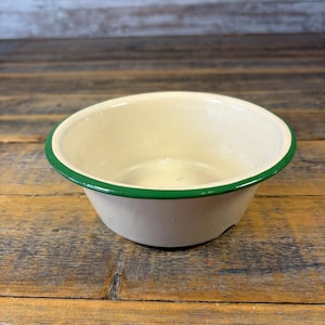May include: Off-white enamel bowl with a bright green rim. The bowl is round and sits on a wooden surface. The interior of the bowl is a creamy off-white color.