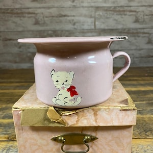 May include: A vintage pink enamel potty chair with a handle and a flared rim. The potty features a hand-painted illustration of a white cat with a red bow. It sits atop a pink box with a brass handle.