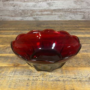 Puede incluir: Un cuenco de vidrio rojo rubí con borde festoneado. El cuenco tiene una base facetada y un color rico y profundo. El vidrio refleja la luz, creando reflejos y sombras. El cuenco es probablemente vintage y adecuado para servir o exhibir.