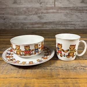 May include: A white ceramic bowl, saucer, and mug set. Each piece features a repeating pattern of brown teddy bears holding hands, with gold trim. The bears wear various outfits in red, blue, and yellow.