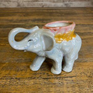 May include: A gray ceramic elephant planter with a pink and yellow flower design on the top. The elephant has a raised trunk and is standing on four legs.