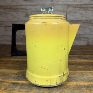 Puede incluir: Cafetera amarilla vintage con asa negra y tapa de cristal. La cafetera tiene un diseño clásico con boquilla y cuerpo cilíndrico. La pintura muestra signos de desgaste, lo que aumenta su encanto antiguo. Un artículo de cocina funcional.