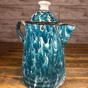 May include: Vintage teal and white enamelware coffee pot with a swirl pattern. The pot has a spout, handle, and a white knob on the lid. The pot is sitting on a wooden surface.