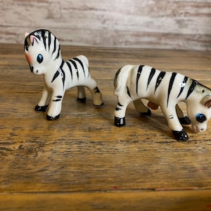 May include: Two ceramic zebra figurines, one standing and one lying down, both with black and white stripes and blue eyes.