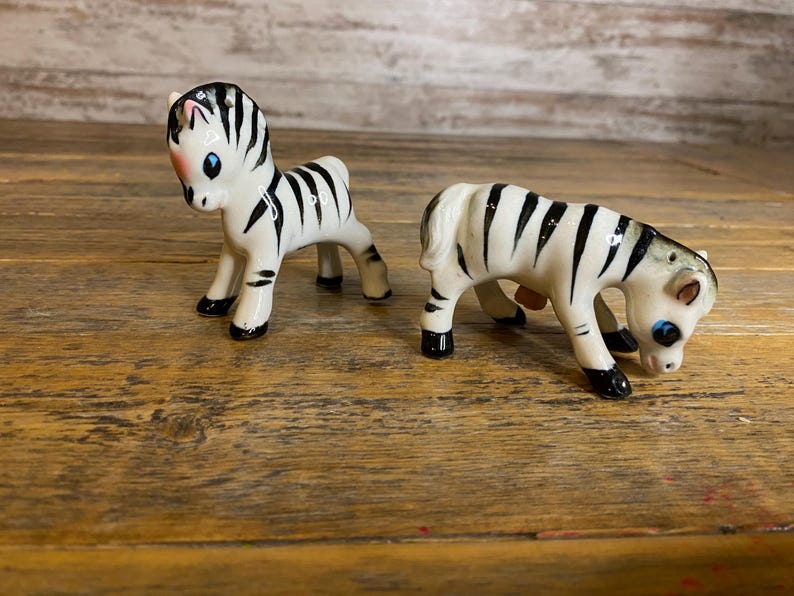 May include: Two ceramic zebra figurines, one standing and one lying down, both with black and white stripes and blue eyes.