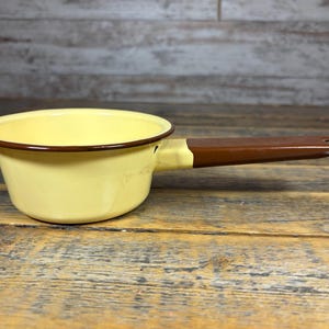 May include: A vintage yellow enamel saucepan with a brown handle. The small saucepan has a rounded shape and a dark brown rim. The handle is long and has a hole at the end for hanging. The pan is set on a wooden surface.