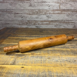 May include: A wooden rolling pin with a natural finish, lying on a rustic wooden surface. The rolling pin has a cylindrical body and handles on each end. The wood grain is visible, adding to its vintage appeal. Suitable for baking.