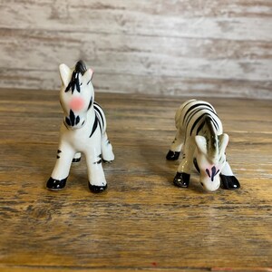 May include: Two ceramic zebra figurines, one standing and one lying down, both with black and white stripes and pink accents on their faces.