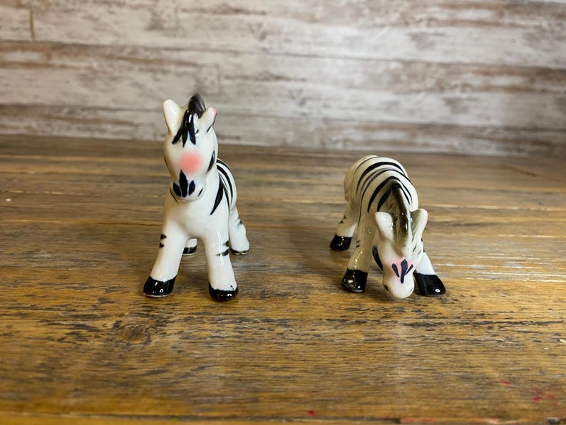 May include: Two ceramic zebra figurines, one standing and one lying down, both with black and white stripes and pink accents on their faces.